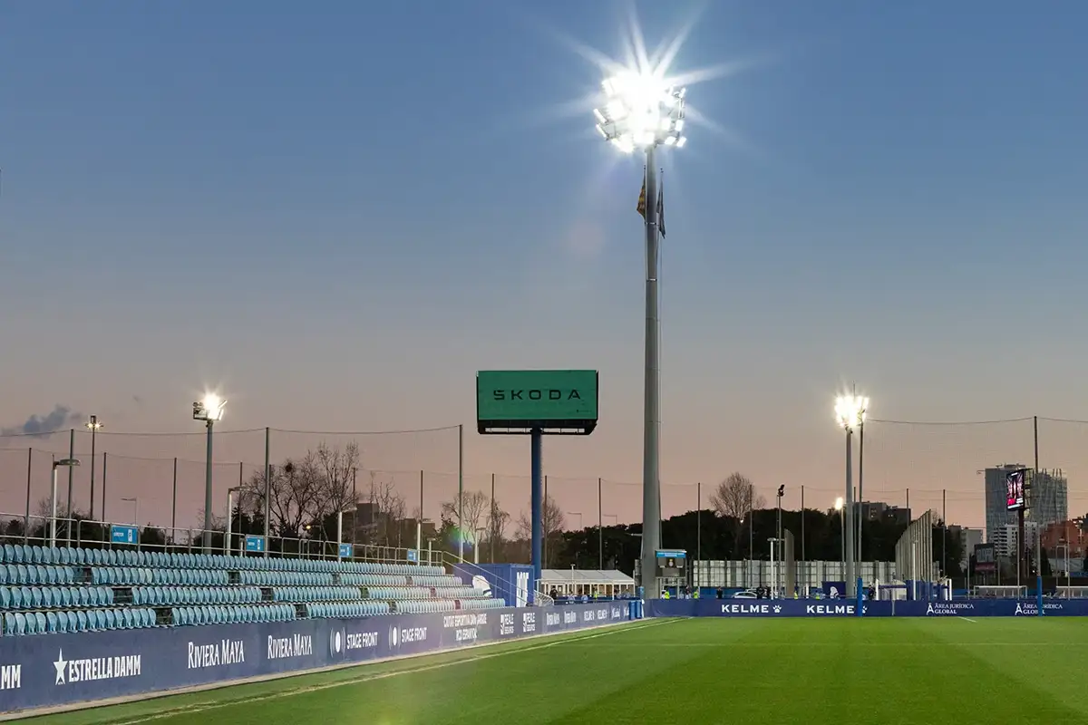 Fotografía de la grada principal iluminada con panel publicitario y focos encendidos al anochecer
