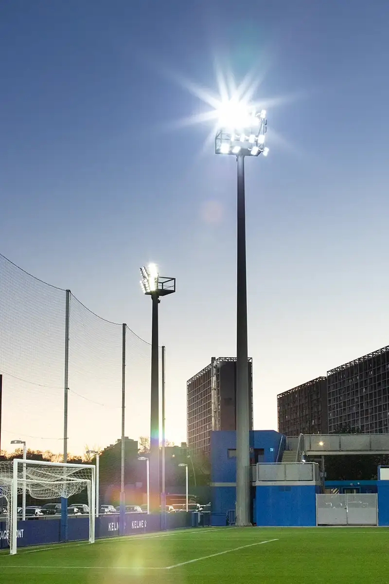 Fotografía de porterías y torres de luz en una esquina del campo con edificios de fondo
