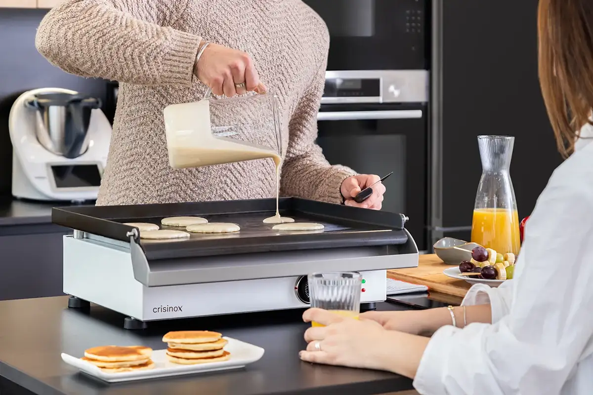 Hombre vertiendo masa sobre una plancha Crisinox mientras comparte desayuno en una cocina moderna