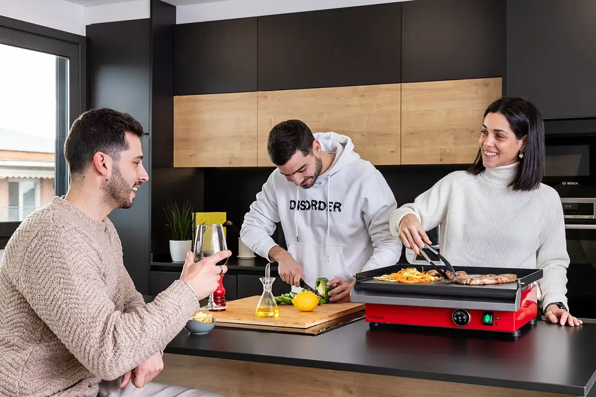 Grupo de amigos cocinando y conversando alrededor de una plancha Crisinox roja en una cocina moderna