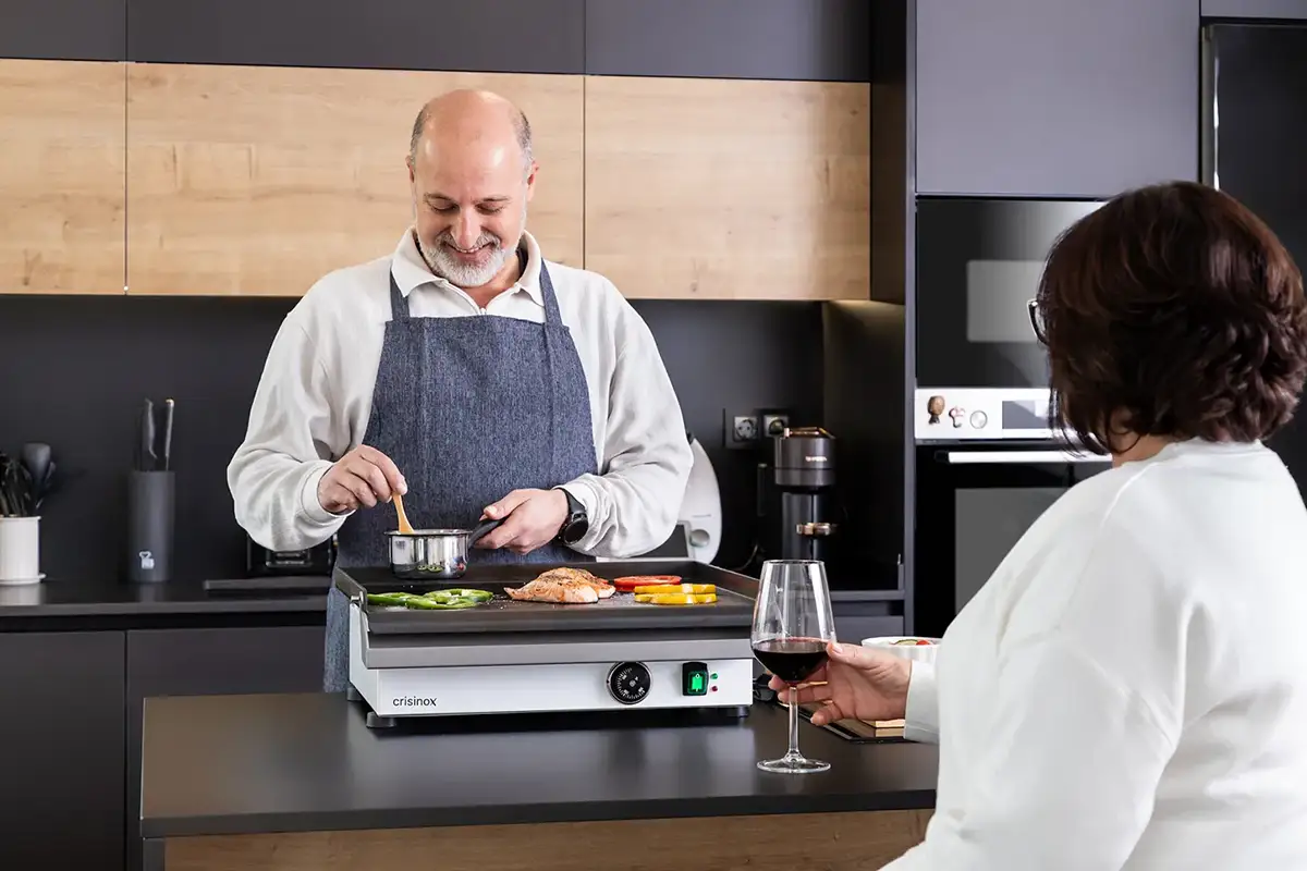 Hombre cocinando en una plancha Crisinox mientras comparte la experiencia con una invitada