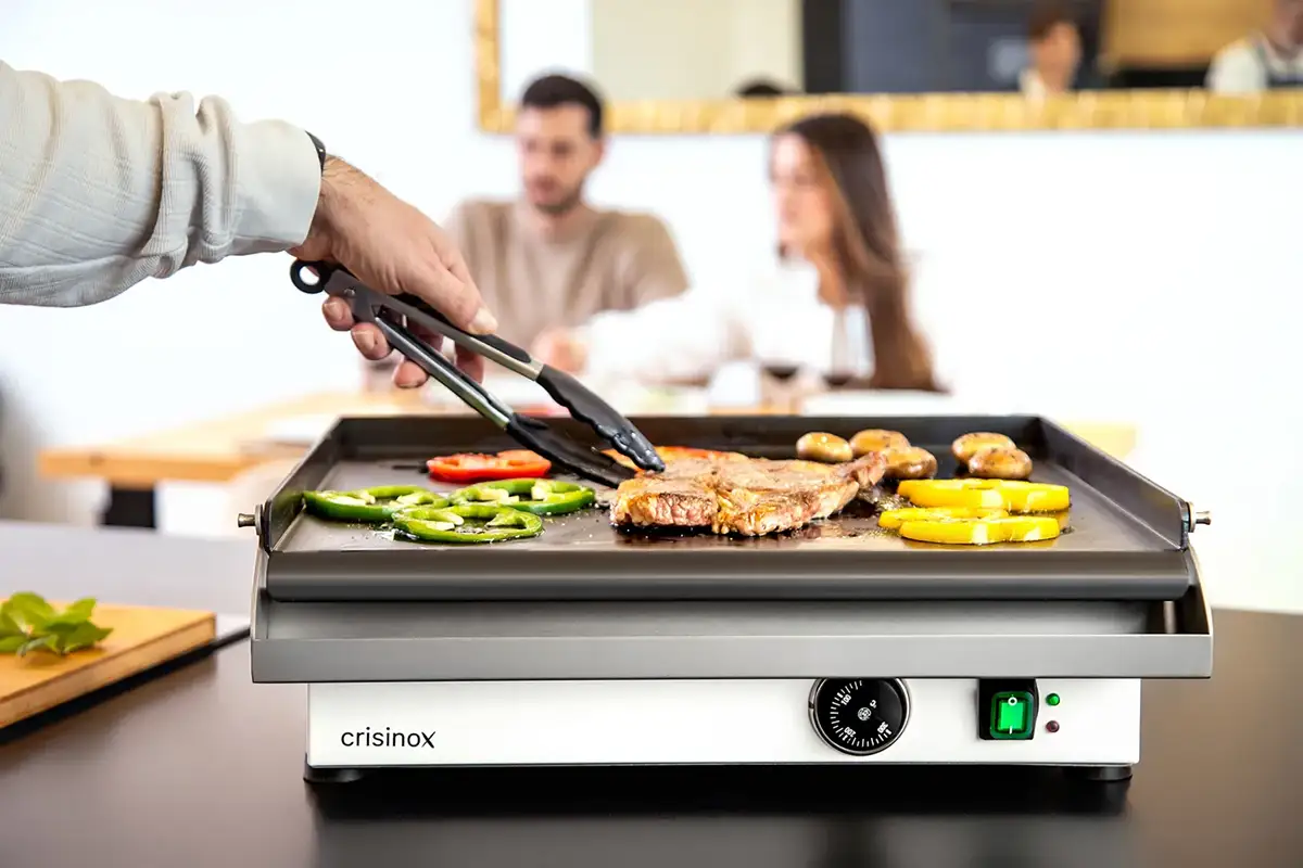 Cocinado de verduras y carne en una plancha Crisinox con comensales al fondo