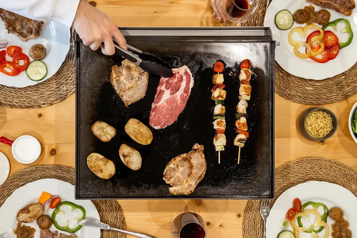 Vista cenital de una plancha Crisinox con carne, patatas y brochetas en medio de una mesa compartida