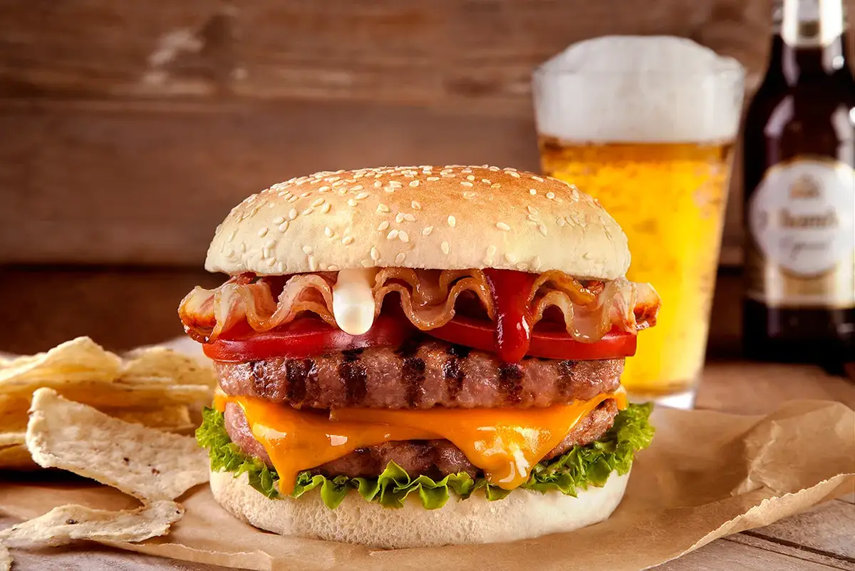 Composición fotográfica de Hamburguesa con queso, bacon y salsa servida con cerveza al fondo sobre mesa de madera