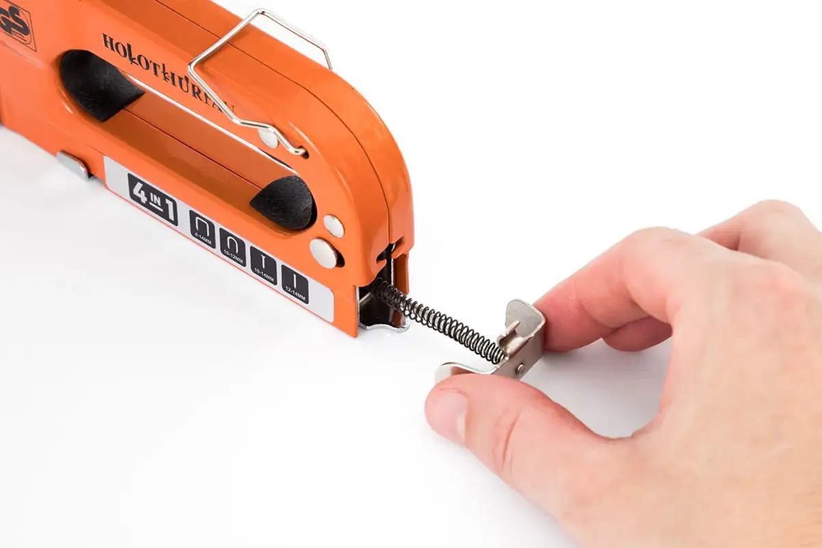 Fotografía de una mano retirando la varilla con resorte del compartimento trasero de la grapadora de mano para cargar grapas para Amazon